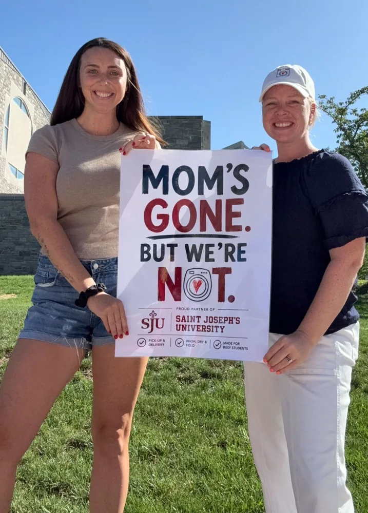 MC and Ally, founders of Lessen the Loads, holding a sign that reads Mom's Gone, But We're Not, proud partner of Saint Joseph's University