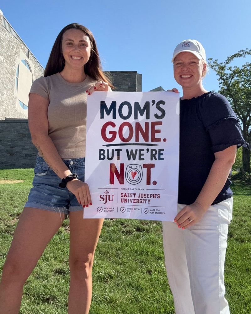 MC and Ally, founders of Lessen the Loads, holding a sign that reads Mom's Gone, But We're Not, proud partner of Saint Joseph's University