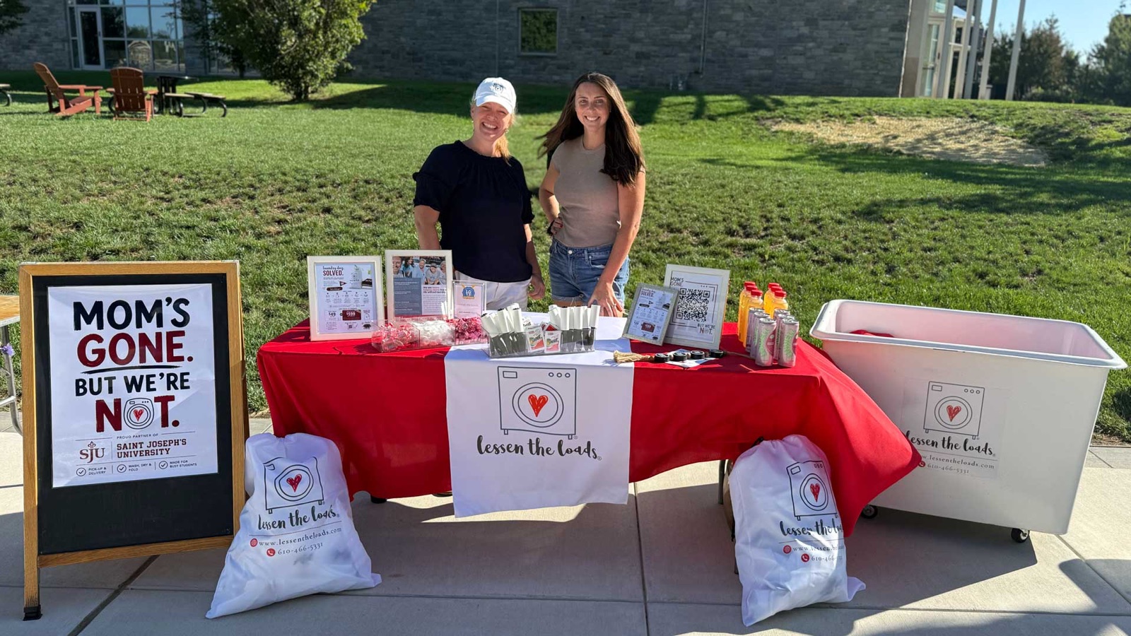 MC and Ally of Lessen the Loads at their table on the Saint Joseph's University campus, with branded laundry bags and a sign reading Mom's Gone, But We're Not